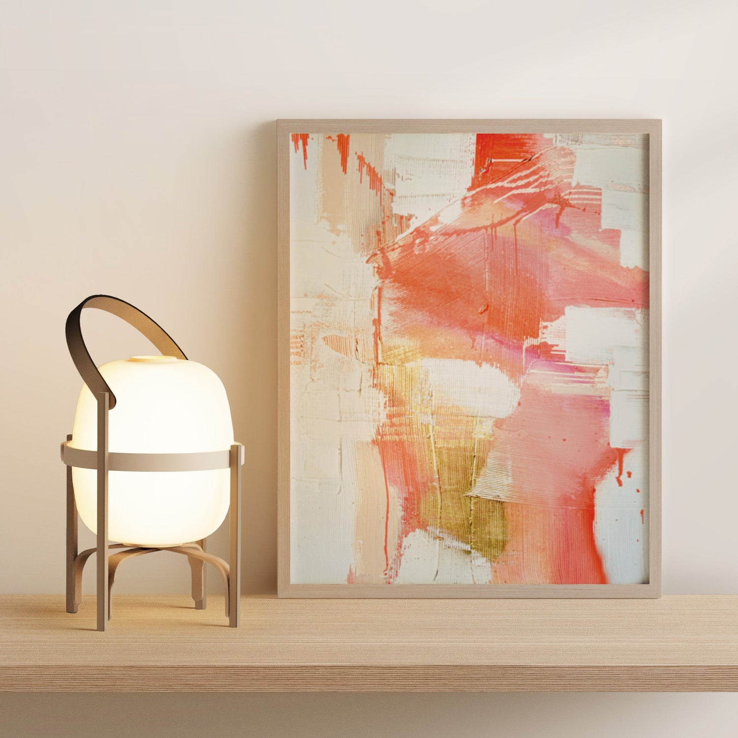 A framed abstract wall art print with coral, pink, orange, and cream brushstrokes leaning against a light wall on a wooden shelf beside a glowing table lamp.